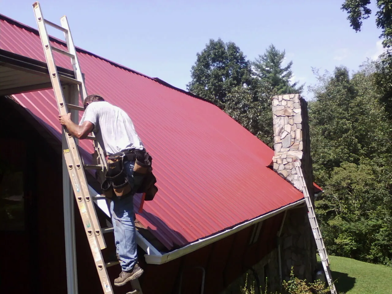 Roofer on a ladder beside a red metal roof and stone chimney — Soffit & Fascia Repair by Roofing Brothers in Duvall